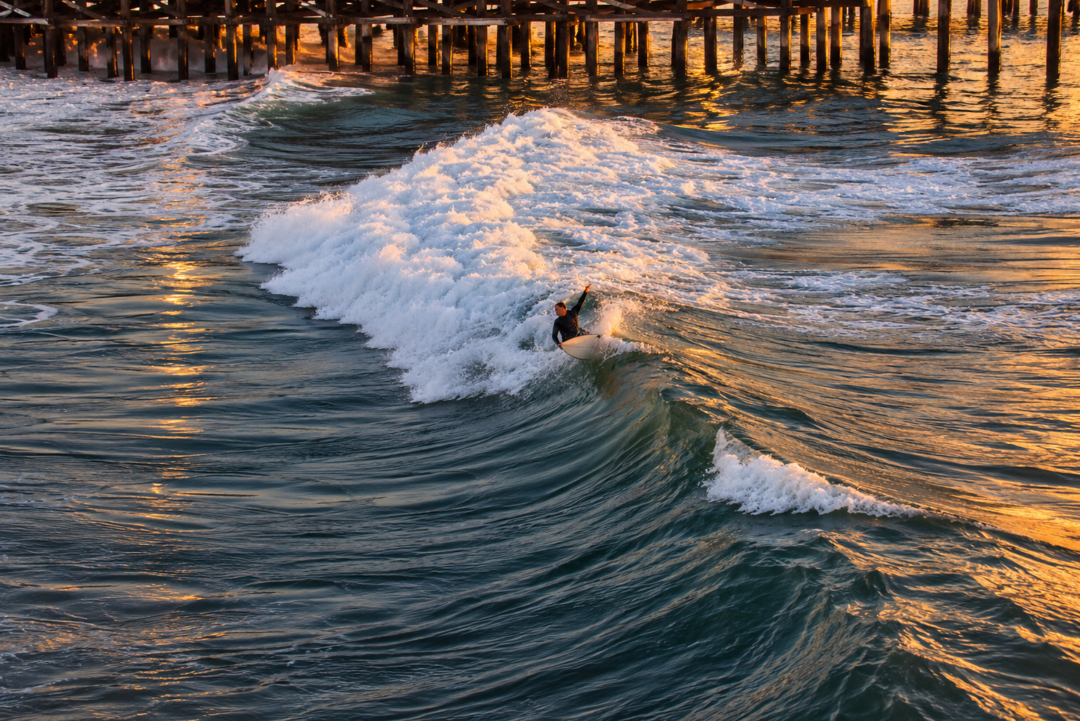 A surfer rides a breaking wave beneath Redondo Beach Pier in golden sunset light